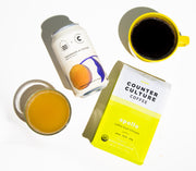 A can and glass of kombucha and a yellow mug of coffee and a bag of Apollo coffee. Overhead shot on a white background.